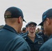 A group of U.S. Navy sailors in dark blue working uniforms are gathered together, with one female sailor wearing glasses and a name tag reading 'PRICE' visible in the center foreground. The sailors are wearing caps with unit designations including 'DDG 113' and 'SFAF' visible on the back of caps in the foreground. The scene appears to be an outdoor formation or informal gathering involving multiple personnel.