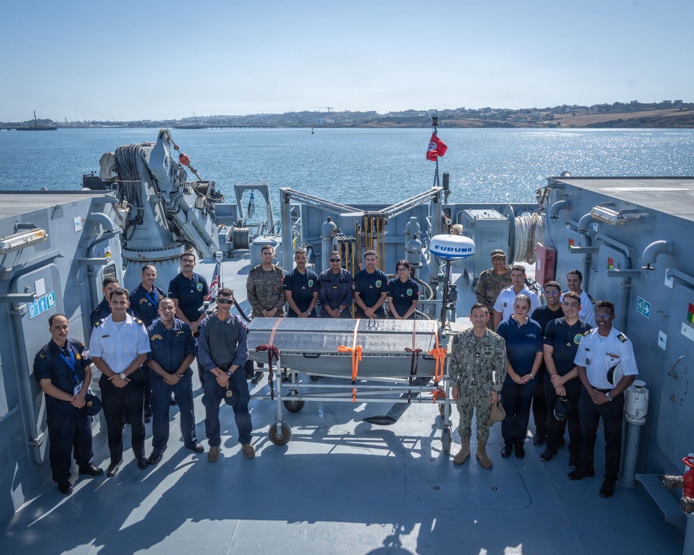 A group of approximately 20 military and civilian personnel are posed for a group photograph on the deck of a naval vessel, likely during a multinational exercise or cooperative event. They are gathered around what appears to be an unmanned underwater vehicle (UUV) or similar maritime equipment on a wheeled cart. The setting is a harbor with calm blue water and a coastal town visible in the background.