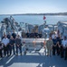 A group of approximately 20 military and civilian personnel are posed for a group photograph on the deck of a naval vessel, likely during a multinational exercise or cooperative event. They are gathered around what appears to be an unmanned underwater vehicle (UUV) or similar maritime equipment on a wheeled cart. The setting is a harbor with calm blue water and a coastal town visible in the background.