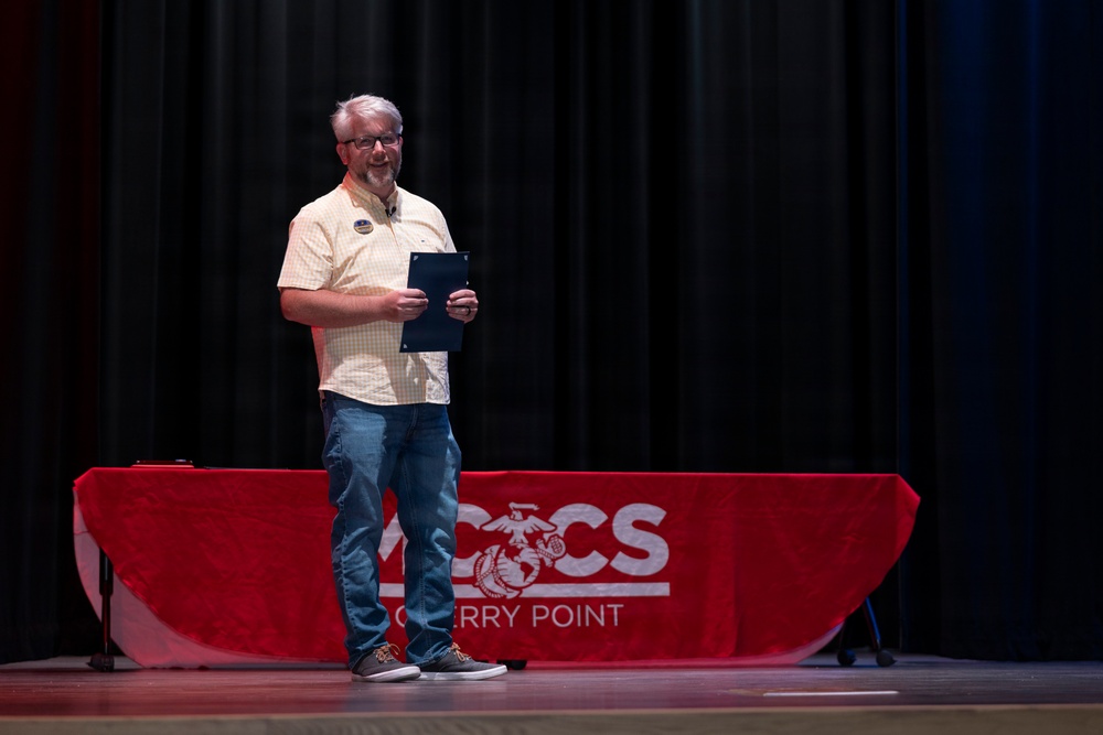 A middle-aged man with gray hair and glasses stands on a stage holding a dark folder or certificate holder. He is positioned in front of a red-draped table bearing the MCCS (Marine Corps Community Services) Cherry Point logo. The setting appears to be a formal or semi-formal presentation or award ceremony.