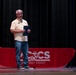 A middle-aged man with gray hair and glasses stands on a stage holding a dark folder or certificate holder. He is positioned in front of a red-draped table bearing the MCCS (Marine Corps Community Services) Cherry Point logo. The setting appears to be a formal or semi-formal presentation or award ceremony.