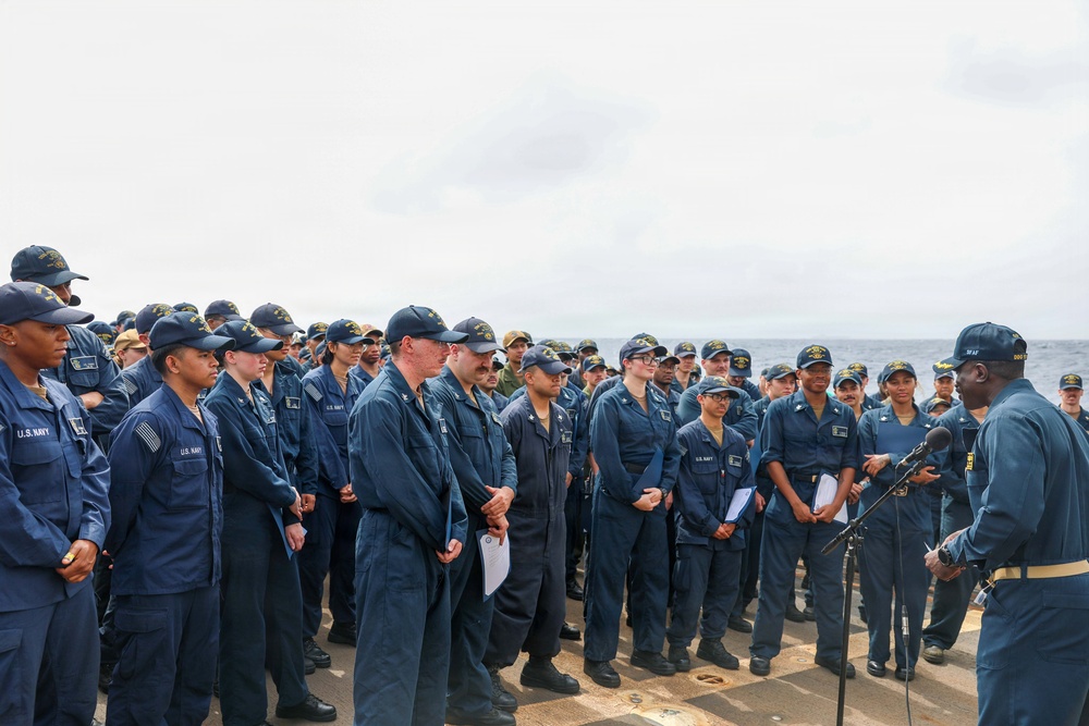 A large group of U.S. Navy personnel assembled on what appears to be the deck of a naval vessel at sea. A senior officer or official is addressing the assembled sailors at a microphone stand on the right side of the image. The personnel are wearing standard Navy working uniforms (NWUs) and cover caps.