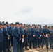 A large group of U.S. Navy personnel assembled on what appears to be the deck of a naval vessel at sea. A senior officer or official is addressing the assembled sailors at a microphone stand on the right side of the image. The personnel are wearing standard Navy working uniforms (NWUs) and cover caps.