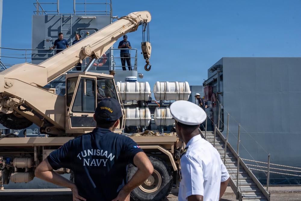The image shows a military logistics operation at a port or dock, where a tan/desert-colored crane vehicle is being used to load or unload equipment near a naval vessel. Two personnel in the foreground are observing the operation — one wearing a 'Tunisian Navy' shirt and another in a white naval officer uniform. Additional personnel are visible on the ship's deck overseeing the crane operation.