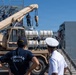 The image shows a military logistics operation at a port or dock, where a tan/desert-colored crane vehicle is being used to load or unload equipment near a naval vessel. Two personnel in the foreground are observing the operation — one wearing a 'Tunisian Navy' shirt and another in a white naval officer uniform. Additional personnel are visible on the ship's deck overseeing the crane operation.