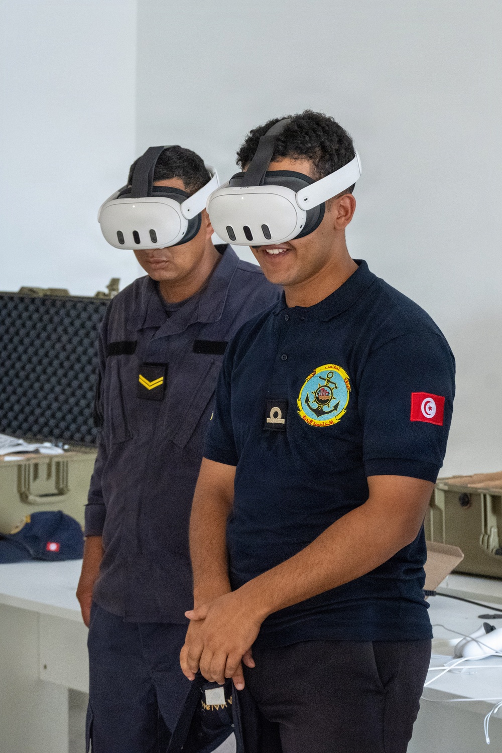 Two military personnel wearing Meta Quest 3 VR headsets are engaged in a virtual reality training or demonstration session. The individual in the foreground wears a dark navy polo shirt bearing a Tunisian Navy emblem and a Tunisian flag patch on the sleeve. Both individuals appear to be actively experiencing VR content in what appears to be an indoor facility.