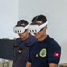 Two military personnel wearing Meta Quest 3 VR headsets are engaged in a virtual reality training or demonstration session. The individual in the foreground wears a dark navy polo shirt bearing a Tunisian Navy emblem and a Tunisian flag patch on the sleeve. Both individuals appear to be actively experiencing VR content in what appears to be an indoor facility.