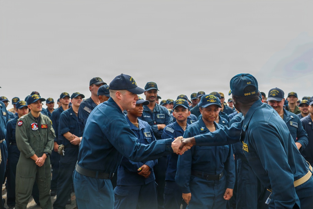 A formal military ceremony aboard what appears to be a U.S. Navy vessel, showing two naval personnel in blue coverall uniforms shaking hands in front of a large assembled crew. The individual on the right wears a cap marked 'CO DDG 113', indicating they are the Commanding Officer of USS John Finn (DDG-113). The assembled crew members are wearing standard Navy working uniforms and caps, with at least one person in an olive/green flight suit visible on the left.