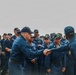 A formal military ceremony aboard what appears to be a U.S. Navy vessel, showing two naval personnel in blue coverall uniforms shaking hands in front of a large assembled crew. The individual on the right wears a cap marked 'CO DDG 113', indicating they are the Commanding Officer of USS John Finn (DDG-113). The assembled crew members are wearing standard Navy working uniforms and caps, with at least one person in an olive/green flight suit visible on the left.