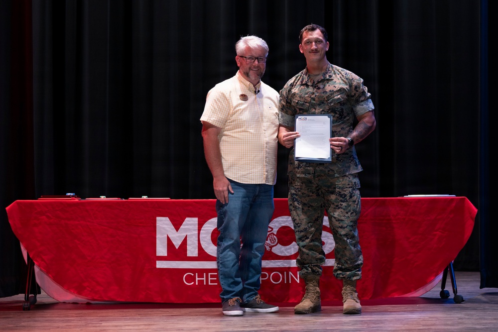 Two men are posing together on a stage in front of a red-draped table bearing the 'MCCS' (Marine Corps Community Services) logo and the word 'Checkpoint.' One man is in civilian clothing while the other is wearing U.S. Marine Corps MARPAT camouflage utilities, and they are jointly holding a document or certificate. The setting appears to be a formal recognition or award ceremony.