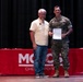 Two men are posing together on a stage in front of a red-draped table bearing the 'MCCS' (Marine Corps Community Services) logo and the word 'Checkpoint.' One man is in civilian clothing while the other is wearing U.S. Marine Corps MARPAT camouflage utilities, and they are jointly holding a document or certificate. The setting appears to be a formal recognition or award ceremony.