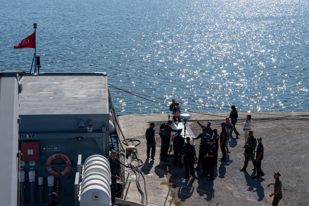 An aerial view of a naval or port facility showing a group of approximately 15-20 personnel gathered on a concrete pier or dock beside a vessel. A Tunisian flag (red with white crescent and star) is visible on a flagpole attached to the ship's structure, and what appears to be a US flag is also present near the group. The scene appears to depict a military or official maritime meeting, demonstration, or equipment inspection.