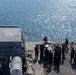 An aerial view of a naval or port facility showing a group of approximately 15-20 personnel gathered on a concrete pier or dock beside a vessel. A Tunisian flag (red with white crescent and star) is visible on a flagpole attached to the ship's structure, and what appears to be a US flag is also present near the group. The scene appears to depict a military or official maritime meeting, demonstration, or equipment inspection.