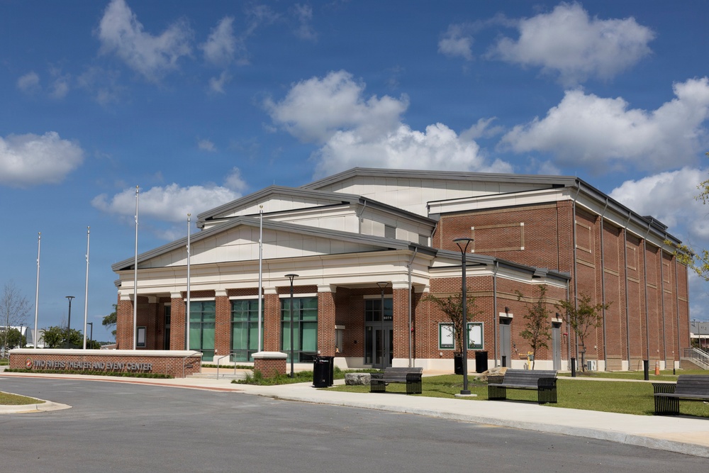 The image shows a modern civic or performing arts building identified as a 'Theater and Event Center,' featuring a combination of brick construction and classical architectural elements including columns and a portico entrance. The facility appears newly constructed or well-maintained, situated on a clean, landscaped campus with parking areas and outdoor seating. The photo was taken on a clear, partly cloudy day with excellent lighting conditions.