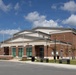 The image shows a modern civic or performing arts building identified as a 'Theater and Event Center,' featuring a combination of brick construction and classical architectural elements including columns and a portico entrance. The facility appears newly constructed or well-maintained, situated on a clean, landscaped campus with parking areas and outdoor seating. The photo was taken on a clear, partly cloudy day with excellent lighting conditions.