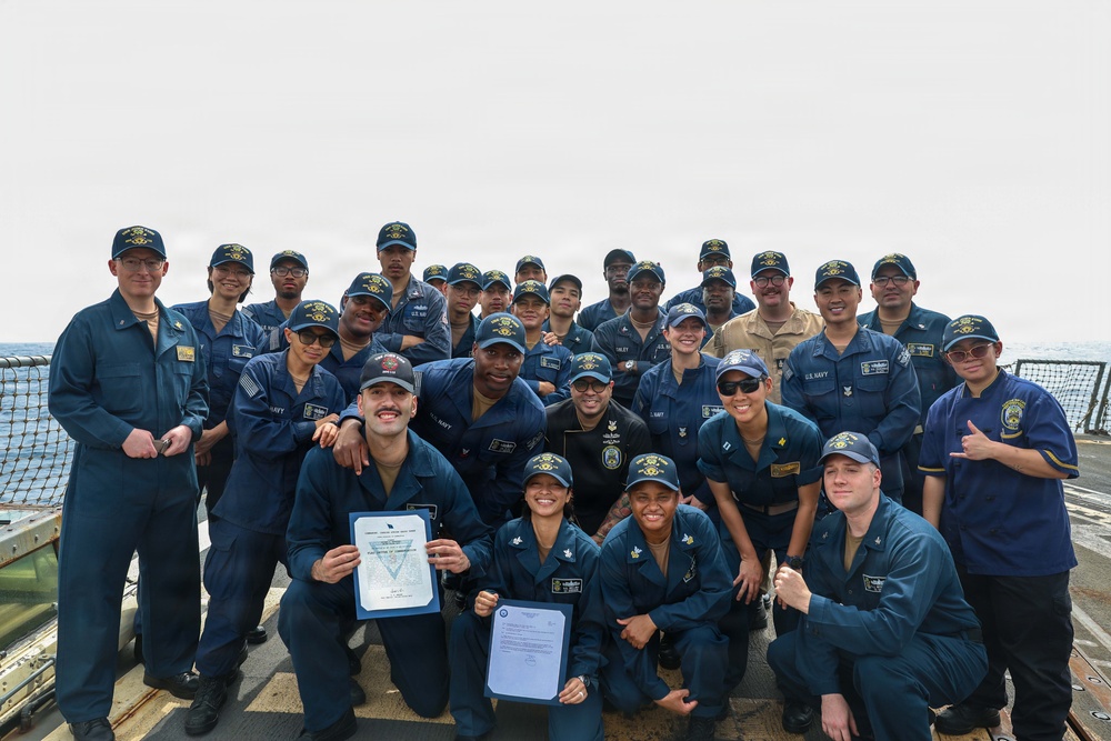 A large group of U.S. Navy personnel pose for a formal group photograph on the deck of a naval vessel at sea. Two individuals in the front row are holding official documents, likely certificates or commendations, suggesting a recognition or award ceremony. The group consists of approximately 25-30 sailors in navy blue coveralls and matching caps.