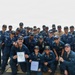 A large group of U.S. Navy personnel pose for a formal group photograph on the deck of a naval vessel at sea. Two individuals in the front row are holding official documents, likely certificates or commendations, suggesting a recognition or award ceremony. The group consists of approximately 25-30 sailors in navy blue coveralls and matching caps.