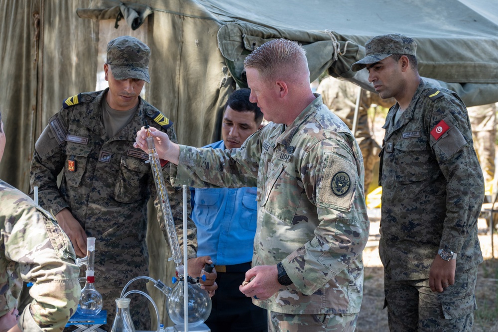 A U.S. Army soldier in MultiCam/OCP camouflage is conducting a demonstration or training exercise involving laboratory glassware and equipment with foreign military personnel. The soldiers on the left and right wear uniforms bearing what appears to be a Tunisian flag patch, suggesting this is a joint military training exercise. The setting is an outdoor field environment with a military tent in the background.