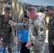 A U.S. Army soldier in MultiCam/OCP camouflage is conducting a demonstration or training exercise involving laboratory glassware and equipment with foreign military personnel. The soldiers on the left and right wear uniforms bearing what appears to be a Tunisian flag patch, suggesting this is a joint military training exercise. The setting is an outdoor field environment with a military tent in the background.