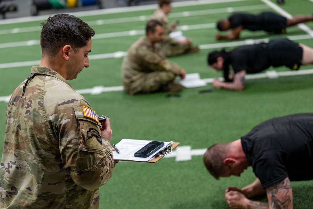 A U.S. Army soldier in MultiCam/OCP uniform with a Ranger tab and American flag patch oversees a physical fitness test on an indoor turf field. He holds a clipboard with documents and a stopwatch, monitoring personnel in black PT uniforms performing what appears to be a push-up or plank exercise. Additional uniformed soldiers are seated in the background, likely serving as graders or awaiting their turn.