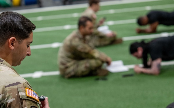 A U.S. Army soldier in MultiCam/OCP uniform with a Ranger tab and American flag patch oversees a physical fitness test on an indoor turf field. He holds a clipboard with documents and a stopwatch, monitoring personnel in black PT uniforms performing what appears to be a push-up or plank exercise. Additional uniformed soldiers are seated in the background, likely serving as graders or awaiting their turn.