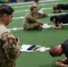 A U.S. Army soldier in MultiCam/OCP uniform with a Ranger tab and American flag patch oversees a physical fitness test on an indoor turf field. He holds a clipboard with documents and a stopwatch, monitoring personnel in black PT uniforms performing what appears to be a push-up or plank exercise. Additional uniformed soldiers are seated in the background, likely serving as graders or awaiting their turn.