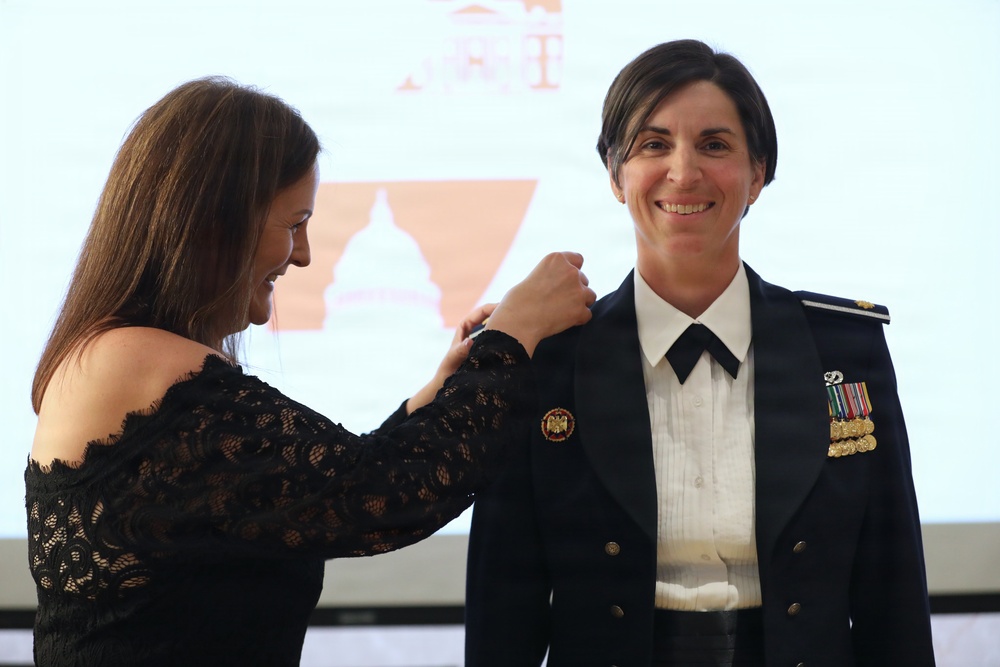 A woman in civilian attire is pinning an insignia or rank device onto the shoulder of a female military officer in dress uniform. The officer is smiling, suggesting this is a formal promotion or award ceremony. A projection screen with a logo or emblem is visible in the background.