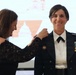 A woman in civilian attire is pinning an insignia or rank device onto the shoulder of a female military officer in dress uniform. The officer is smiling, suggesting this is a formal promotion or award ceremony. A projection screen with a logo or emblem is visible in the background.