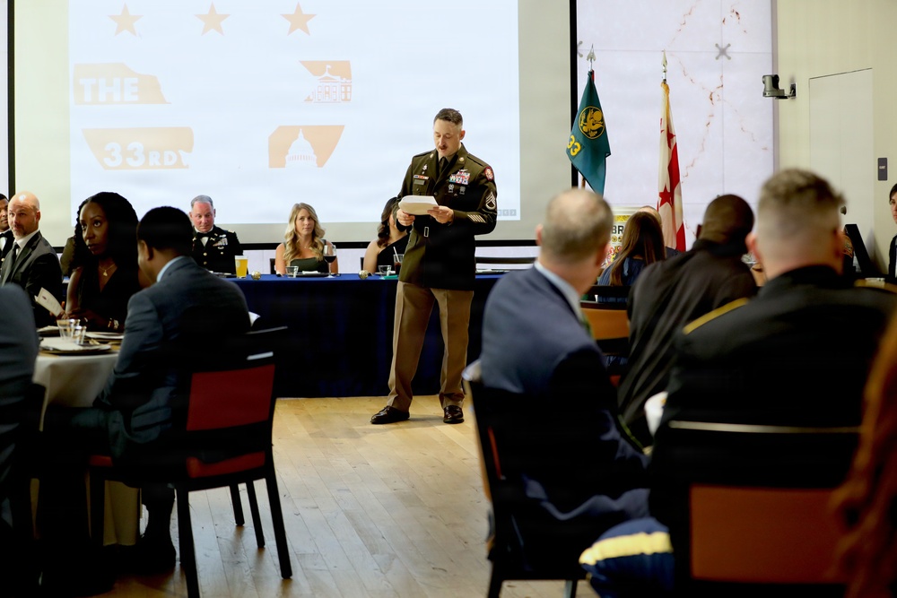 A formal military banquet or ceremony is taking place in a large hall, with a uniformed soldier standing at the center reading from a document or paper. A projection screen behind him displays text reading 'THE 33RD' along with stars and graphic elements, suggesting this is a unit anniversary or recognition event. Attendees in military uniforms and civilian formal attire are seated at round and rectangular tables.