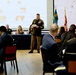 A formal military banquet or ceremony is taking place in a large hall, with a uniformed soldier standing at the center reading from a document or paper. A projection screen behind him displays text reading 'THE 33RD' along with stars and graphic elements, suggesting this is a unit anniversary or recognition event. Attendees in military uniforms and civilian formal attire are seated at round and rectangular tables.