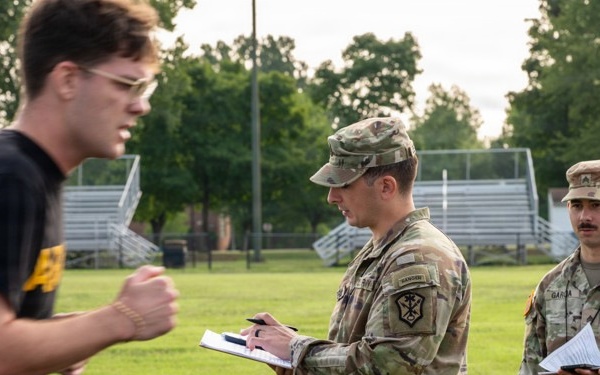 Two U.S. Army soldiers in OCP (Operational Camouflage Pattern) uniforms are observing and recording data on clipboards while a young man in civilian athletic wear (black t-shirt and shorts) appears to be performing a physical fitness test on a grass field. The setting appears to be a military physical fitness assessment, likely an Army Combat Fitness Test (ACFT) or similar evaluation. Bleachers and trees are visible in the background.