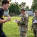 Two U.S. Army soldiers in OCP (Operational Camouflage Pattern) uniforms are observing and recording data on clipboards while a young man in civilian athletic wear (black t-shirt and shorts) appears to be performing a physical fitness test on a grass field. The setting appears to be a military physical fitness assessment, likely an Army Combat Fitness Test (ACFT) or similar evaluation. Bleachers and trees are visible in the background.