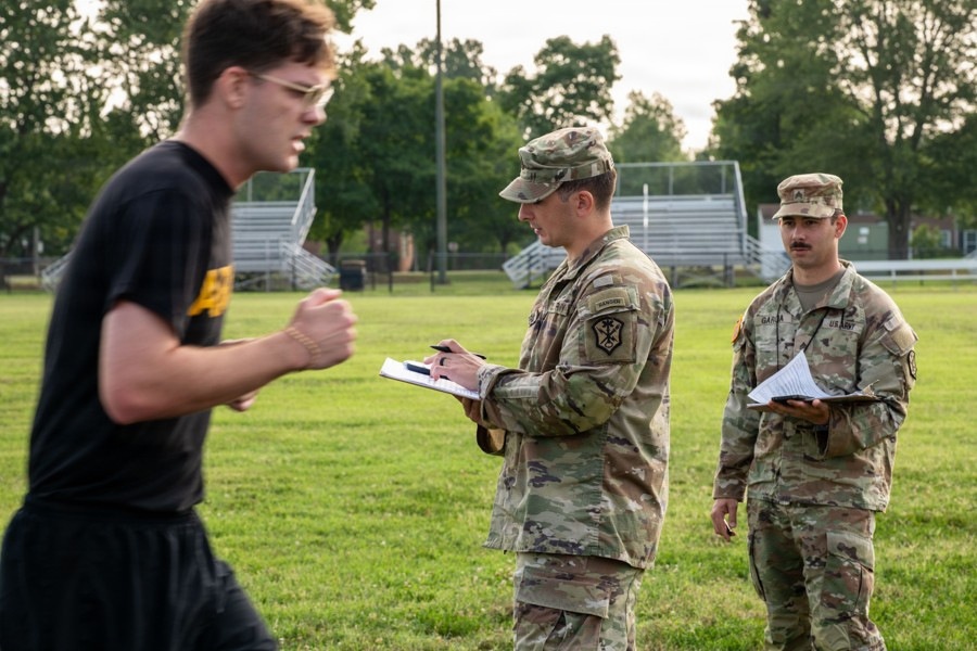 Two U.S. Army soldiers in OCP (Operational Camouflage Pattern) uniforms are observing and recording data on clipboards while a young man in civilian athletic wear (black t-shirt and shorts) appears to be performing a physical fitness test on a grass field. The setting appears to be a military physical fitness assessment, likely an Army Combat Fitness Test (ACFT) or similar evaluation. Bleachers and trees are visible in the background.