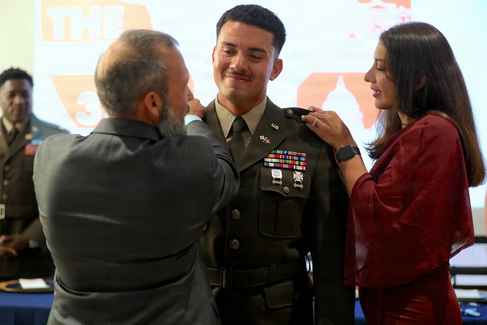 A U.S. Army officer is being pinned with rank or insignia on his shoulders by two individuals — an older man in a gray suit on the left and a young woman in a red outfit on the right. The officer is wearing the Army Service Uniform (ASU) with multiple ribbons, badges, and U.S. collar brass, suggesting this is a promotion or commissioning ceremony. The setting appears to be an indoor military ceremony with a projected presentation visible in the background.