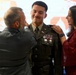 A U.S. Army officer is being pinned with rank or insignia on his shoulders by two individuals — an older man in a gray suit on the left and a young woman in a red outfit on the right. The officer is wearing the Army Service Uniform (ASU) with multiple ribbons, badges, and U.S. collar brass, suggesting this is a promotion or commissioning ceremony. The setting appears to be an indoor military ceremony with a projected presentation visible in the background.