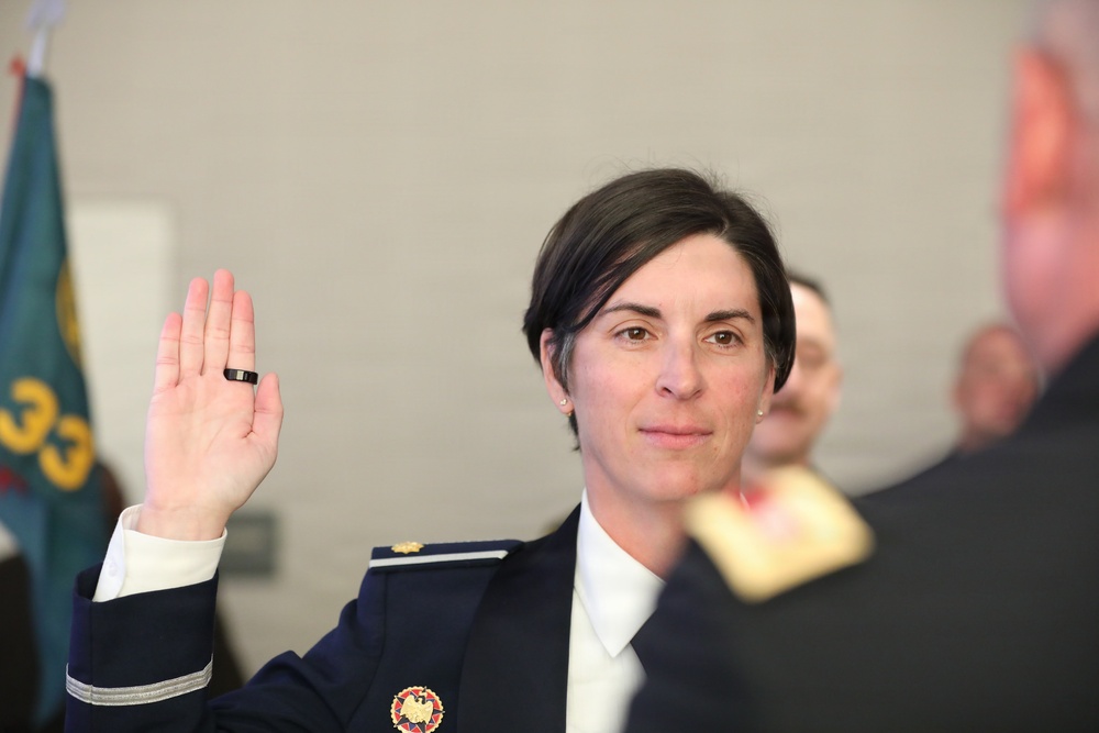 A woman in a dark navy military or law enforcement dress uniform is raising her right hand, appearing to take an oath or be sworn in during a formal ceremony. She is wearing a badge/insignia on her chest and a ring on her finger. Other uniformed individuals are visible in the background, along with what appears to be a flag.