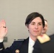 A woman in a dark navy military or law enforcement dress uniform is raising her right hand, appearing to take an oath or be sworn in during a formal ceremony. She is wearing a badge/insignia on her chest and a ring on her finger. Other uniformed individuals are visible in the background, along with what appears to be a flag.