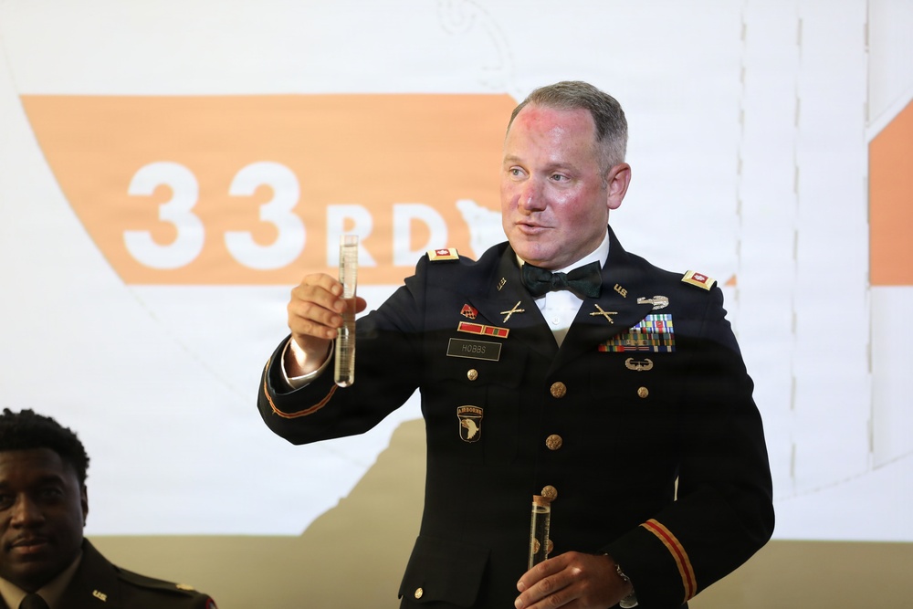 A U.S. Army officer in dress uniform (Army Service Uniform with bow tie, indicating a formal military event) is holding up what appears to be small vials or test tubes. His name tag reads 'HOBBS' and he wears the 101st Airborne Division patch along with multiple service ribbons and crossed rifles insignia. The background displays a projection reading '33RD,' suggesting this is likely a military anniversary or formal dining-in/dining-out ceremony.