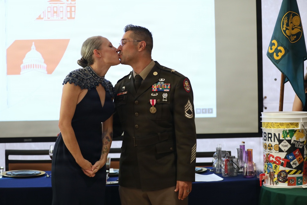 A U.S. Army soldier in dress uniform shares a kiss with a woman in a navy blue formal dress at what appears to be a military formal event or ball. The soldier displays multiple medals, ribbons, and rank insignia consistent with a Staff Sergeant. A green unit flag marked '33' and a CBRN/Hazmat Team bucket are visible in the background.