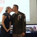 A U.S. Army soldier in dress uniform shares a kiss with a woman in a navy blue formal dress at what appears to be a military formal event or ball. The soldier displays multiple medals, ribbons, and rank insignia consistent with a Staff Sergeant. A green unit flag marked '33' and a CBRN/Hazmat Team bucket are visible in the background.