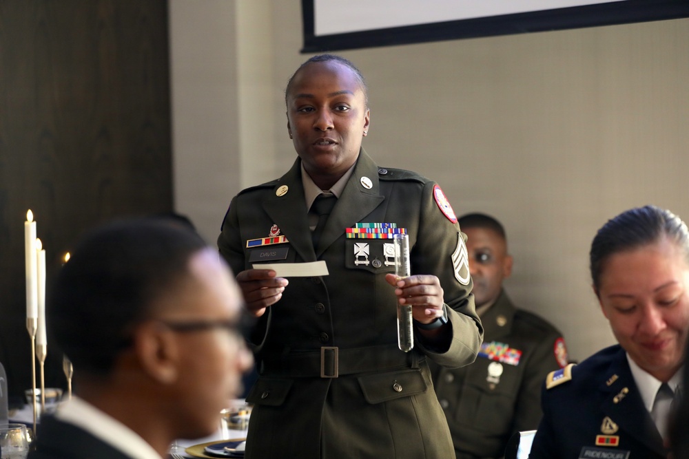 A U.S. Army soldier in dress green uniform, identified by a name tag reading 'DAVIS,' stands and speaks at what appears to be a formal military dining event or ceremony. She holds a glass and a card or note, suggesting she is delivering a toast or reading remarks. Other uniformed military personnel are seated at the table around her.