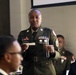 A U.S. Army soldier in dress green uniform, identified by a name tag reading 'DAVIS,' stands and speaks at what appears to be a formal military dining event or ceremony. She holds a glass and a card or note, suggesting she is delivering a toast or reading remarks. Other uniformed military personnel are seated at the table around her.