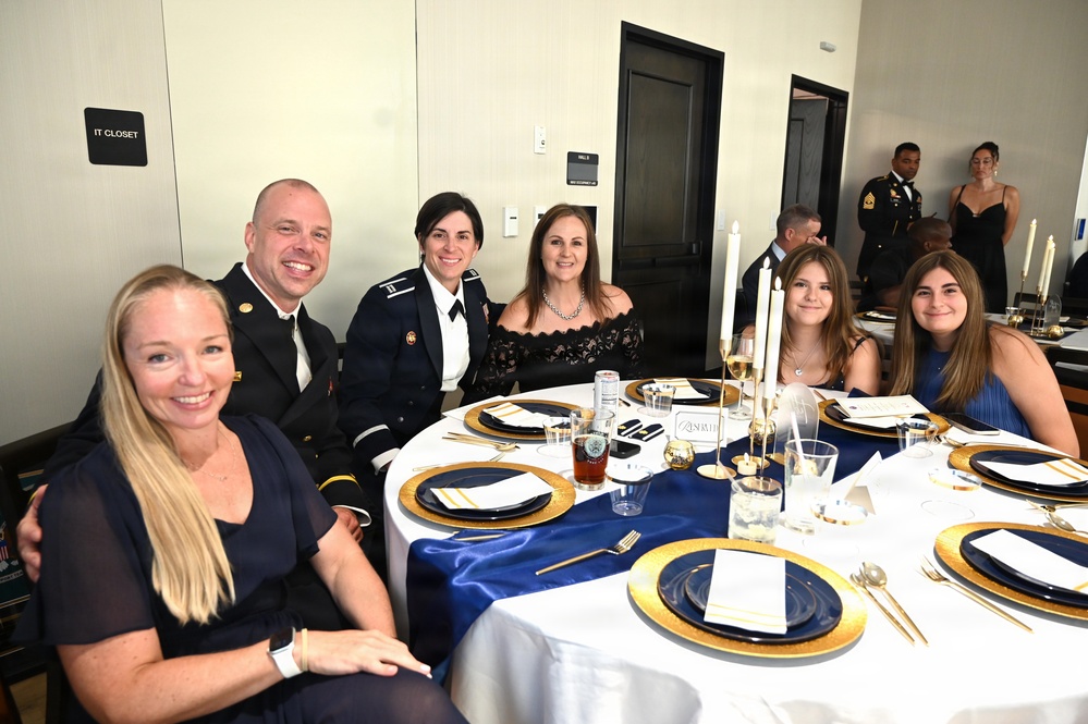 A group of six people are seated at a formally set round banquet table at what appears to be a military formal dinner or ball. Two individuals are wearing U.S. Army dress uniforms, while the others are dressed in formal civilian attire. The table is elegantly decorated with navy blue and gold charger plates, white linens, candles, and glassware.