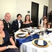 A group of six people are seated at a formally set round banquet table at what appears to be a military formal dinner or ball. Two individuals are wearing U.S. Army dress uniforms, while the others are dressed in formal civilian attire. The table is elegantly decorated with navy blue and gold charger plates, white linens, candles, and glassware.