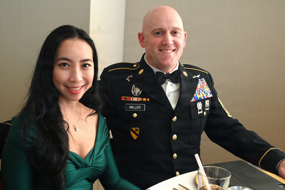 Two individuals are seated at a formal dining table, posing for a photo. The man is wearing a U.S. Army dress uniform (Army Service Uniform - Black) with the name tag 'MILLER,' multiple service ribbons, a 1st Cavalry Division patch, and senior NCO rank insignia. The woman is wearing an elegant green evening dress, suggesting this is a formal military event such as a military ball.