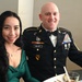 Two individuals are seated at a formal dining table, posing for a photo. The man is wearing a U.S. Army dress uniform (Army Service Uniform - Black) with the name tag 'MILLER,' multiple service ribbons, a 1st Cavalry Division patch, and senior NCO rank insignia. The woman is wearing an elegant green evening dress, suggesting this is a formal military event such as a military ball.