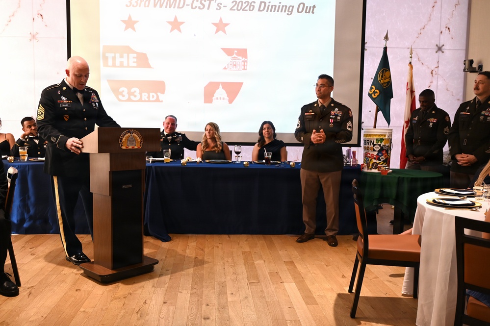 A formal military dining out ceremony for the 33rd WMD-CST (Weapons of Mass Destruction - Civil Support Team) is taking place in 2026. A senior non-commissioned officer, identifiable as a Sergeant Major by rank insignia, is speaking at a podium while another soldier stands at attention nearby. Military personnel in dress uniforms and civilian guests are seated at a head table beneath a presentation screen.