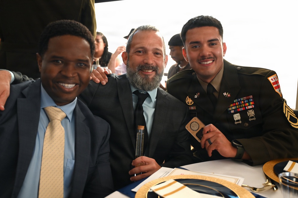 Three men pose together smiling at what appears to be a formal military ceremony or banquet. The man on the right is wearing a U.S. Army dress uniform with multiple service ribbons and rank insignia, while the other two men are dressed in civilian business attire. They appear to be seated at a table and are holding small commemorative items.
