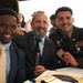 Three men pose together smiling at what appears to be a formal military ceremony or banquet. The man on the right is wearing a U.S. Army dress uniform with multiple service ribbons and rank insignia, while the other two men are dressed in civilian business attire. They appear to be seated at a table and are holding small commemorative items.