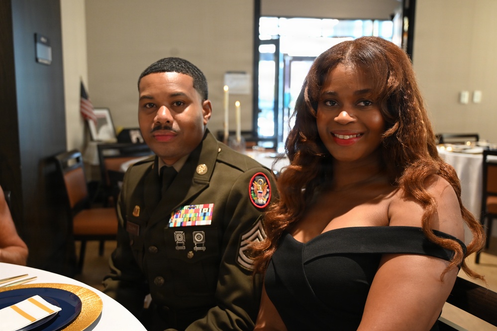 Two individuals are seated at a formal dining table, posing for a photo. The man on the left is wearing a U.S. Army dress uniform with multiple service ribbons, rank insignia consistent with a Senior Non-Commissioned Officer (Master Sergeant or Sergeant First Class), and a U.S. Army shoulder patch. The woman on the right is wearing an elegant off-shoulder black dress, suggesting this is a formal military event or banquet.